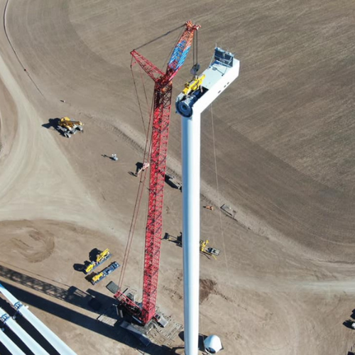 Red crane lifting a large white wind turbine blade on a construction site.