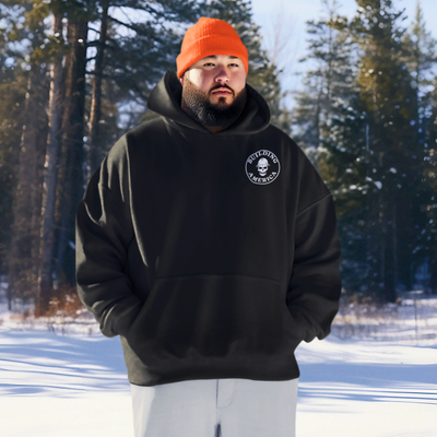 Large bearded man wearing a black bella canvas crane operator hoodie with building America DTG printed on the front with a orange construction hoodie on the woods with snow