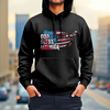 A man wearing a black hoodie with "God Bless America" printed on the front in a distressed American flag design. He is standing in front of a city skyline at sunset.