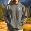 Hispanic male with a gray bella canvas hoodie on with his hands in the kangaroo pouch pocket. The hoodie has a DTG printed logo with building america on it on the front while standing against a mountain and woods background