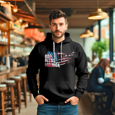 A man with short hair and a beard wearing a black hoodie with "God Bless America" printed on the front in a distressed American flag design. He is standing in a warmly lit café or restaurant interior.