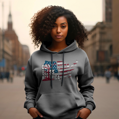 A woman with curly hair wearing a gray hoodie with "God Bless America" printed on the front in a distressed American flag design. She is standing on a city street with buildings visible in the background.